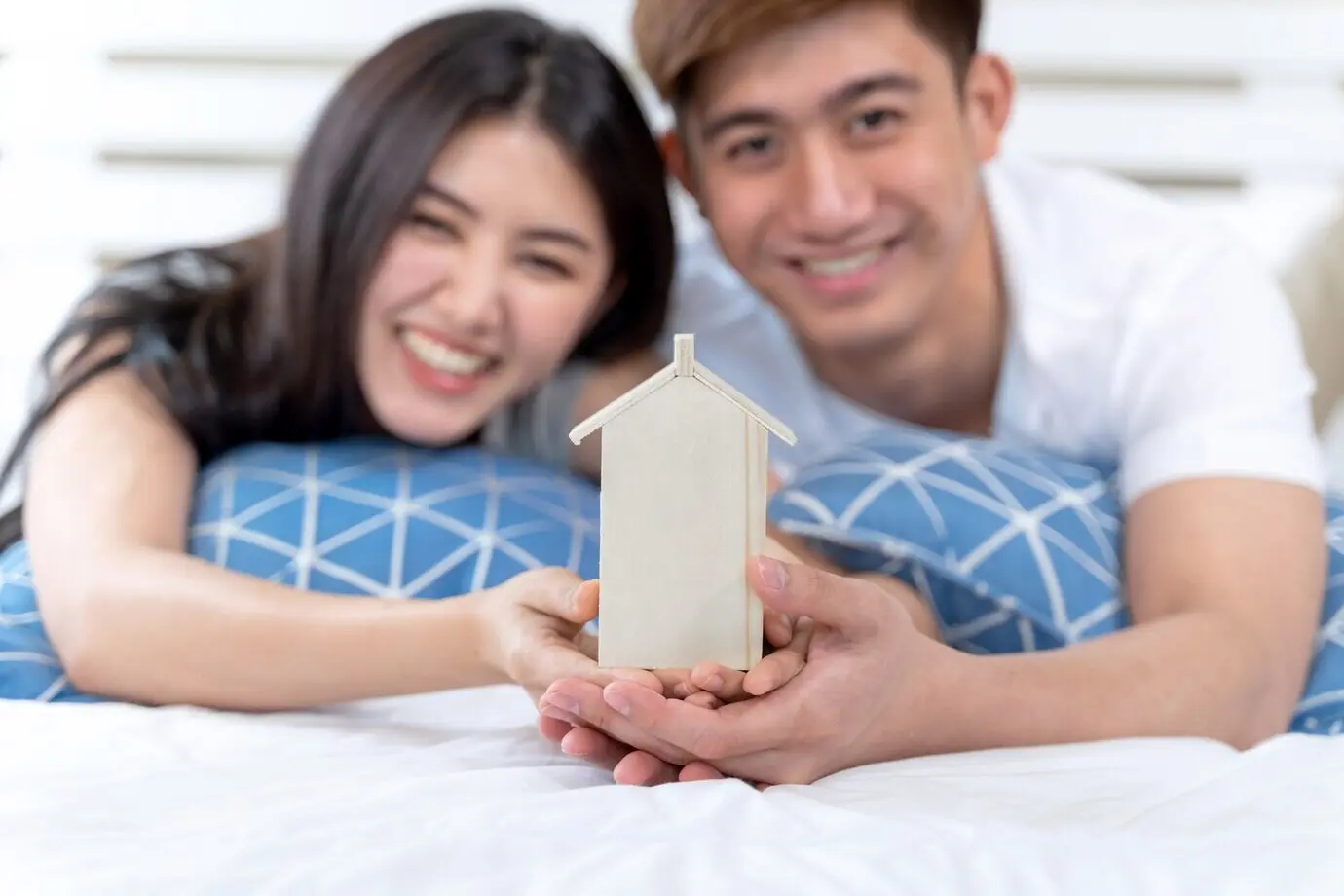 A young couple holding a toy house on the bed.